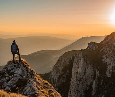 a lone hiker standing on a rocky peak overlooking a breathtaking mountain landscape at sunset with hues of orange and gold 13 adventures in nature 13 peaks to explore 13 moments of tranquility