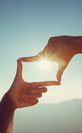 two hands framing a bright sun with a clear blue sky in the background symbolizing perspective and enlightenment on the fifth dimension