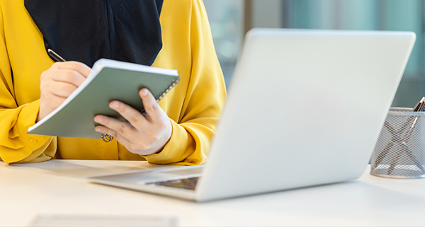 person wearing a yellow blouse taking notes in a notebook while sitting at a desk with a laptop and pen this setup emphasizes productivity and organization ideal for multitasking and efficient planning